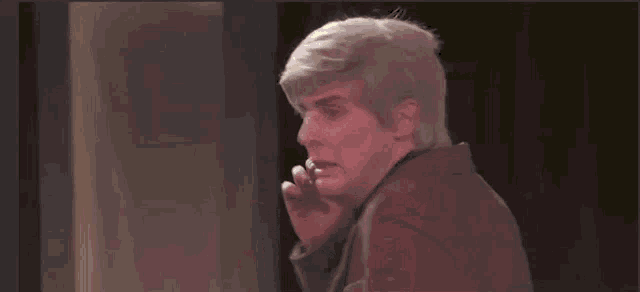 a man in a brown suit and black turtleneck is standing in front of a black wall .