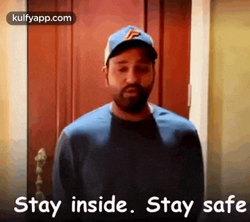 a man wearing a baseball cap is standing in front of a door and says `` stay inside . stay safe '' .