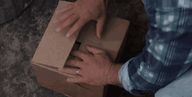 a man in a plaid shirt is opening a cardboard box on the ground .