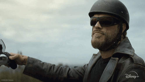 a man with a beard wearing a helmet and sunglasses is riding a motorcycle with a disney logo behind him