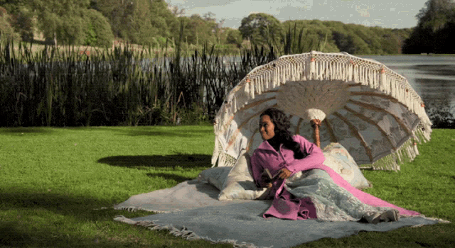 a woman sits on a blanket under an umbrella in the grass