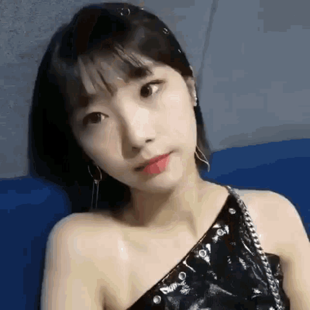 a woman is sitting on a blue couch wearing a black one shoulder top and earrings .