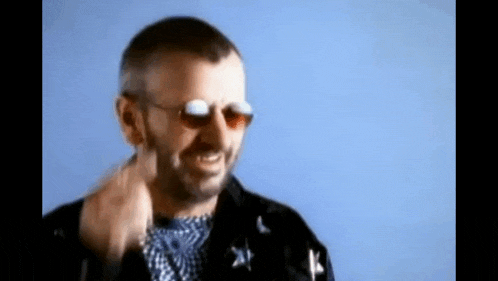 a man wearing sunglasses and a black jacket is smiling and pointing .