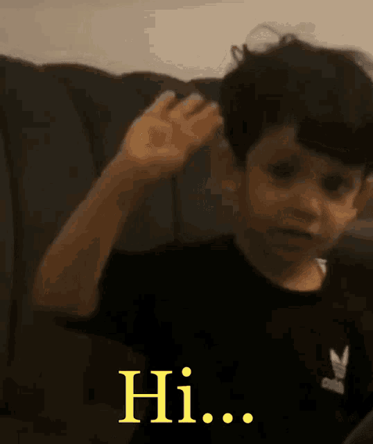 a young boy is sitting on a couch waving his hand and saying hi .