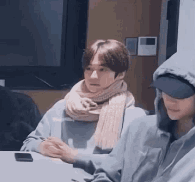 a man wearing a scarf and a hoodie is sitting at a table with another man .