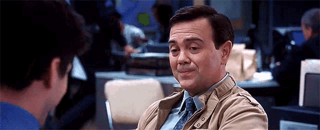 a man in a tan jacket and tie is sitting at a table with another man .