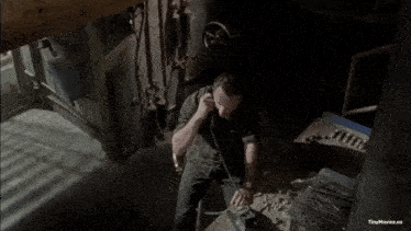 a man in a black shirt is holding a gun in a dark room with tinymoviez.co written on the bottom