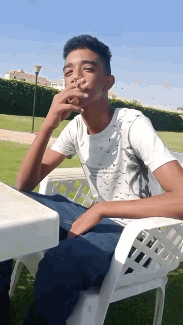 a young man is sitting in a chair smoking a cigarette while wearing a white shirt with birds on it