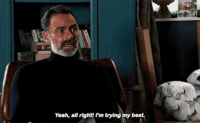 a man with a beard is sitting in a chair and says " yeah all right i 'm trying my best "
