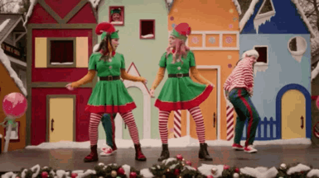 three people dressed as elves are dancing in front of gingerbread houses