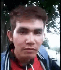 a man wearing a red shirt and a blue jacket is standing in front of a tree and looking at the camera .