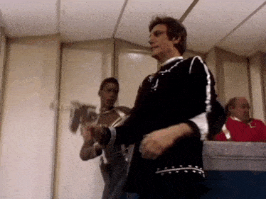 a man in a black sweater is dancing in a room while a man in a red shirt looks on .