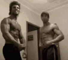 two muscular men are standing next to each other in a room without shirts .