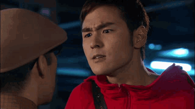a man in a red jacket looks at another man with his eyes closed