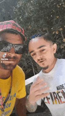 two men are posing for a picture together and one of them is wearing sunglasses and a hat .