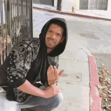 a man in a black hoodie is squatting down on the sidewalk and giving the middle finger