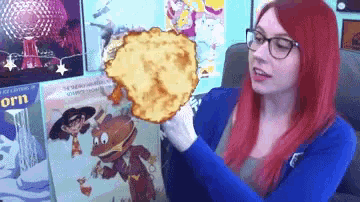 a woman with red hair and glasses holds a cartoon character in front of a poster that says orn