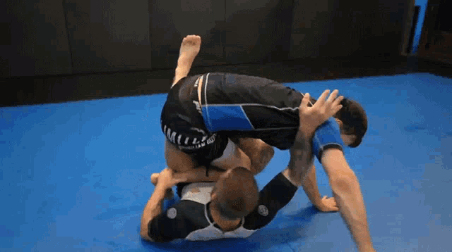two men are wrestling on a blue mat and one of them is wearing a black shirt that says ' sss ' on it