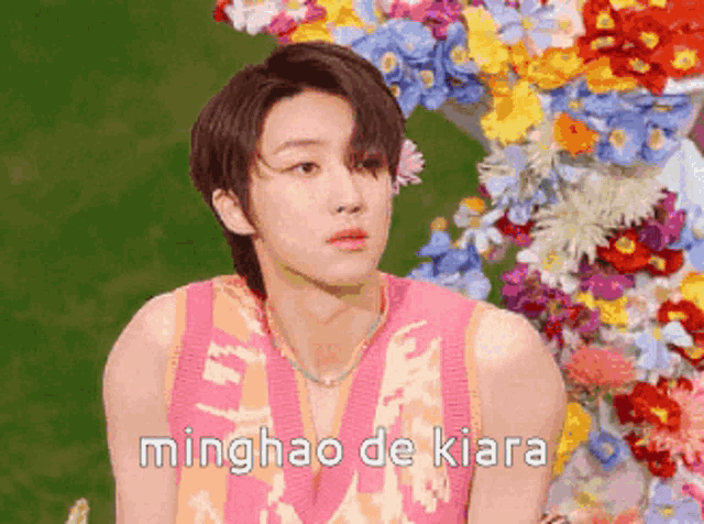 a young man in a pink shirt is sitting in front of a bunch of flowers and says minghao de kiara .