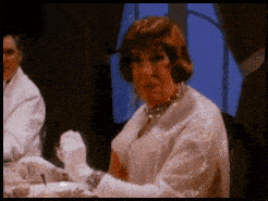 a woman wearing white gloves and a necklace sits at a table