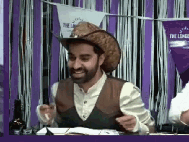 a man wearing a cowboy hat and vest is sitting at a table with his hands in the air .