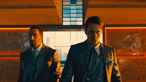 two men in suits standing next to each other in front of a window