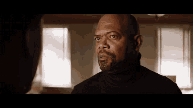 a bald man with a beard wearing a black turtleneck looks at another man