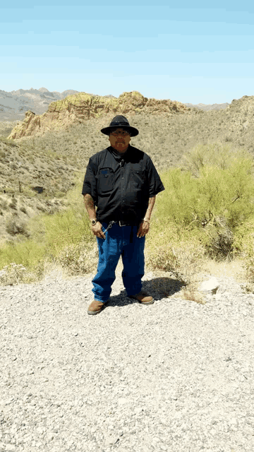 a man wearing a black shirt and a hat stands on a gravel road
