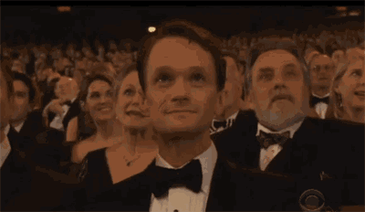 a man in a tuxedo is sitting in a crowd of people looking up at something .
