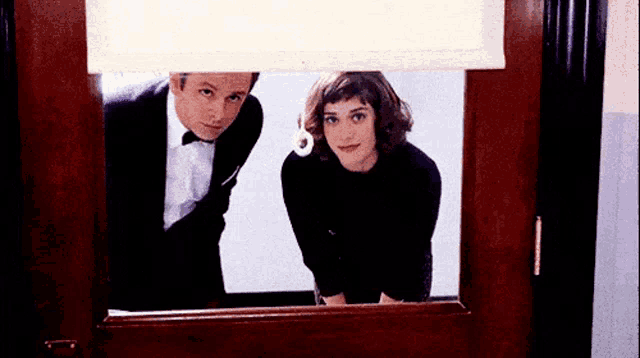 a man in a tuxedo and a woman in a black dress are looking out of a window .