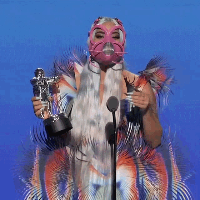 a woman in a pink mask holds a trophy in front of a microphone