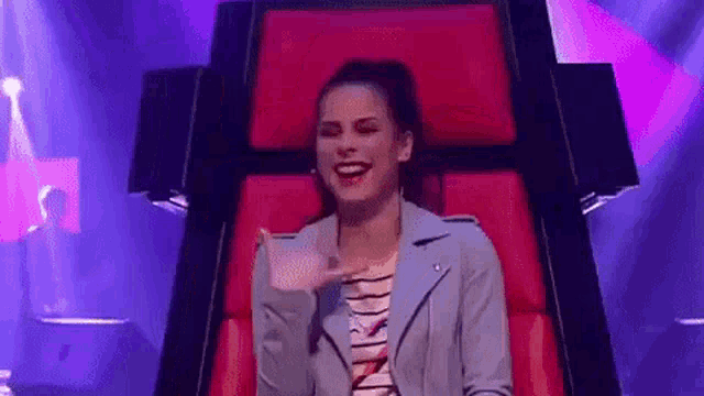 a woman in a blue jacket is sitting in a red chair and laughing .