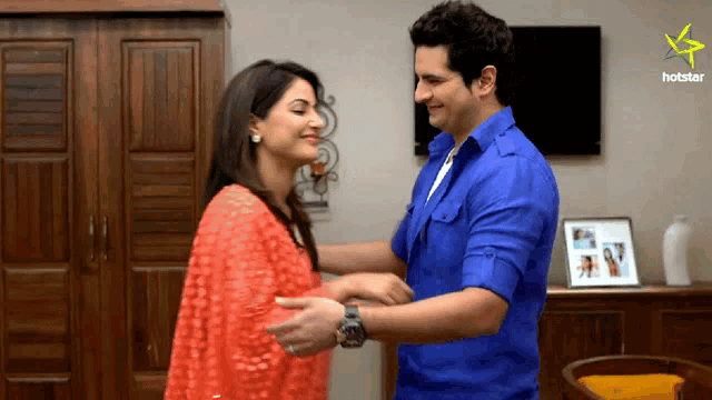 a man in a blue shirt is hugging a woman in a red top in front of a tv screen that says hotstar