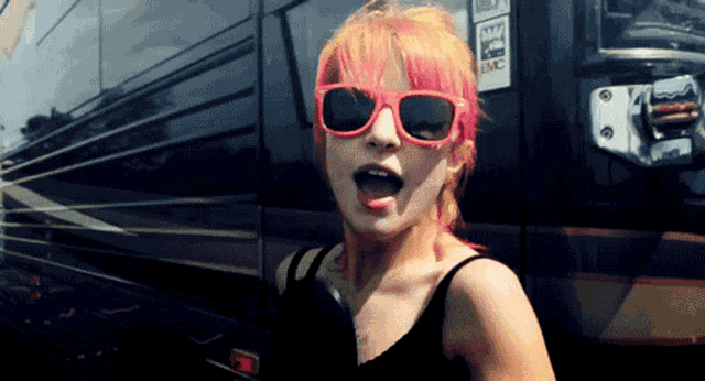 a woman with pink hair wearing sunglasses stands in front of a bus