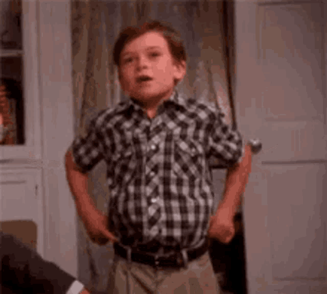 a young boy in a plaid shirt is standing in front of a door and making a funny face .