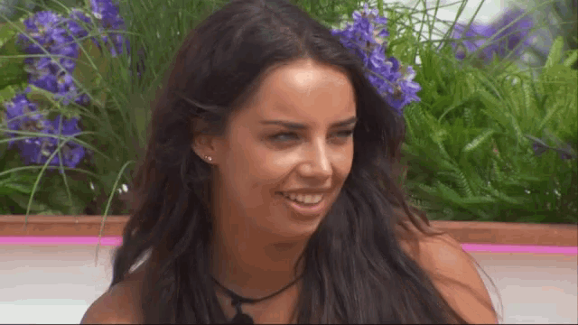 a woman with long dark hair is smiling in front of some purple flowers