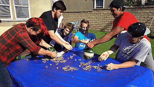 a group of young men are gathered around a table with a blue table cloth and one of them is wearing a shirt that says ma