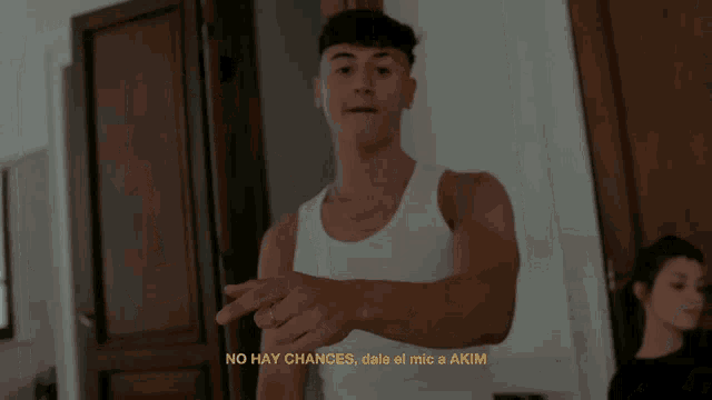 a man in a white tank top is standing in a room with a woman sitting at a table