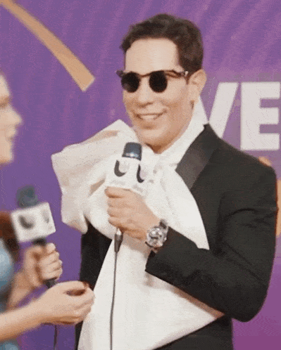 a man wearing sunglasses and a tuxedo is talking into a microphone with the letter e in the background