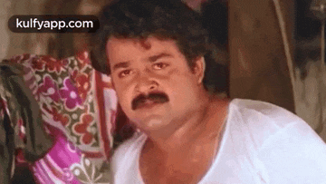 a man with a mustache is sitting on a bed and looking at the camera .