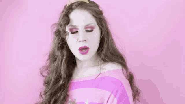 a woman wearing a pink sweater and a tiara is making a funny face against a pink background .