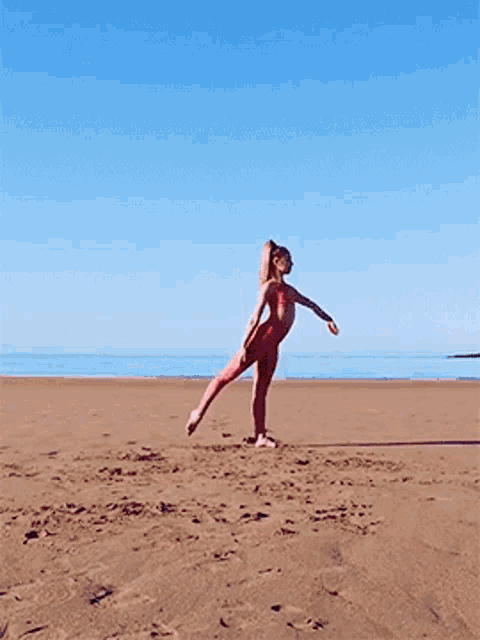 a woman in a red bodysuit stands on one leg on the beach
