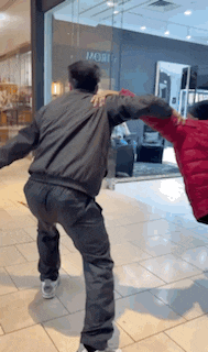a man in a gray jacket is holding another man in a red jacket in a mall .