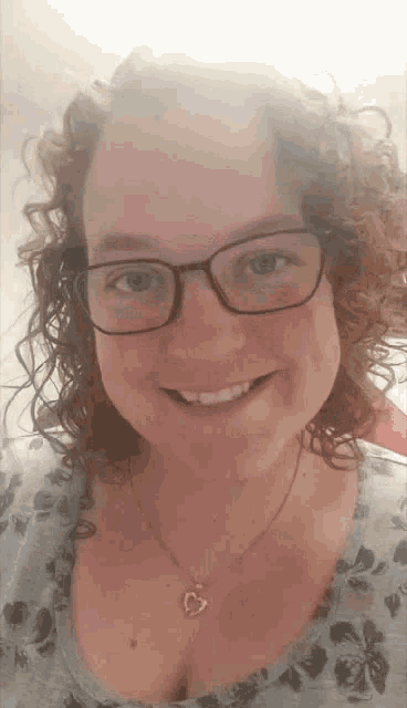 a woman with curly hair wearing glasses and a heart necklace is smiling .