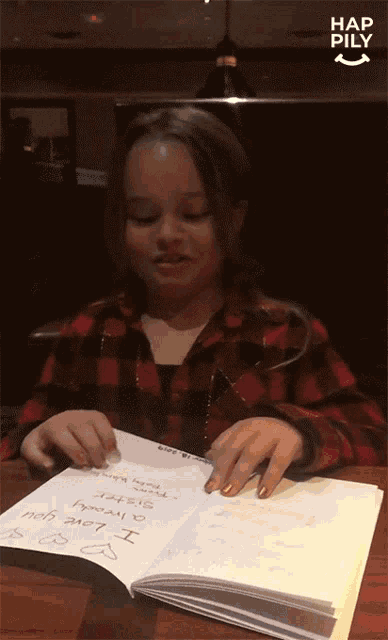 a girl in a plaid shirt is sitting at a table with a book open to a page that says " i love you "