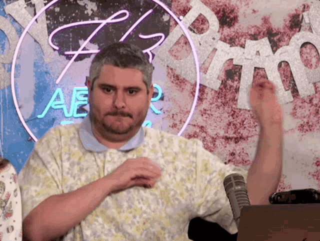 a man with a beard is sitting in front of a microphone in a room with a neon sign behind him .