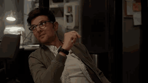 a man wearing glasses and a watch is sitting in a dark room holding a pen .