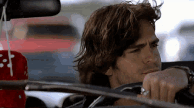 a man with long hair is driving a car and looking at the rear view mirror while wearing a ring on his finger .