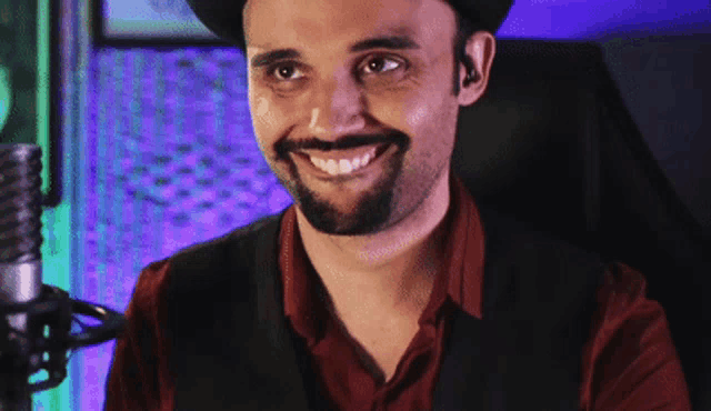 a man with a beard wearing a hat and vest smiles in front of a microphone