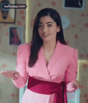 a woman in a pink jacket and red belt is standing in a room with her hands outstretched .